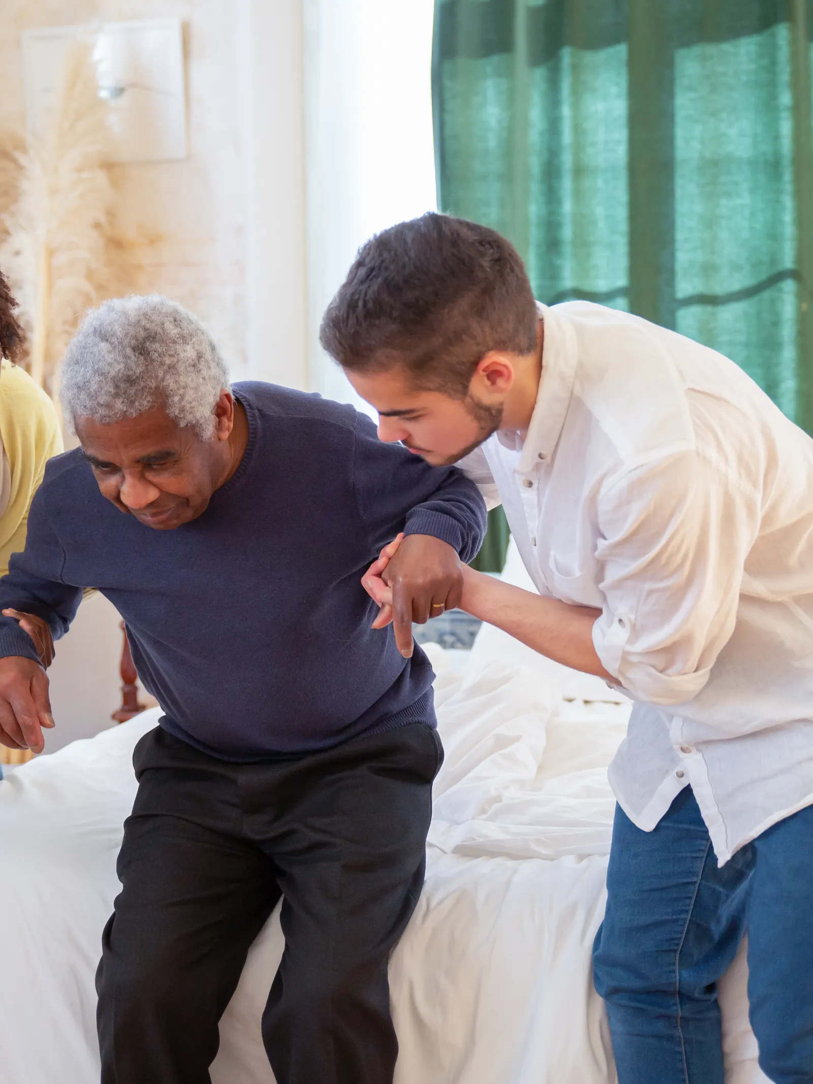 Dois profissionais de home care apoiando um paciente idoso a se levantar da cama, em casa.